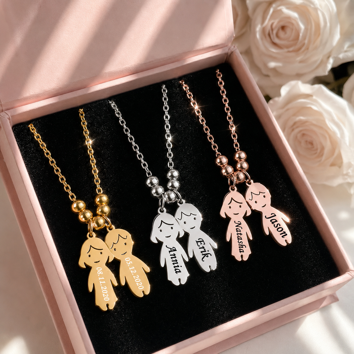 Four personalized necklaces with child-shaped pendants and names on a black box with pink interior, surrounded by white roses.