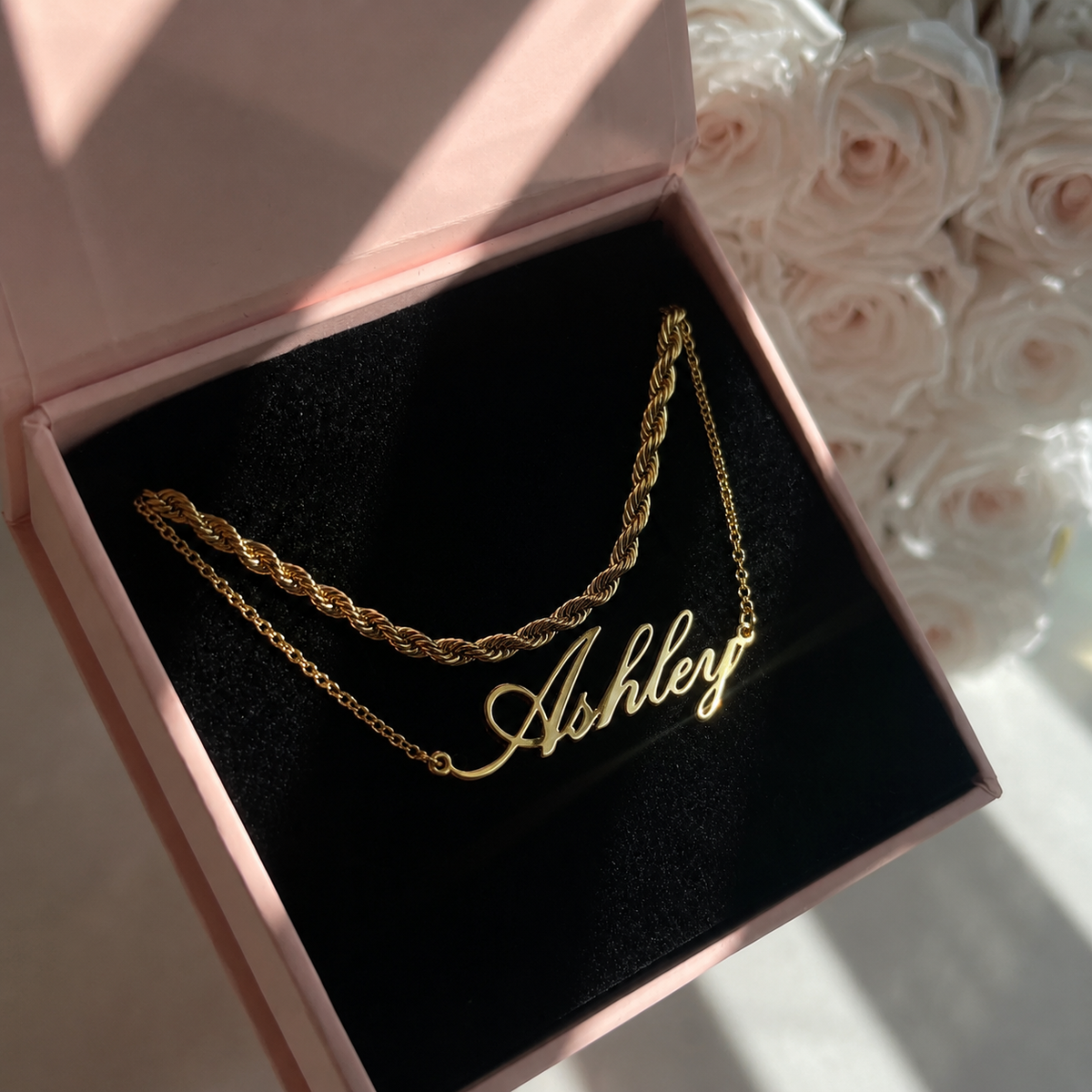 Gold necklace with 'Ashley' engraving in a pink jewelry box with flowers in the background