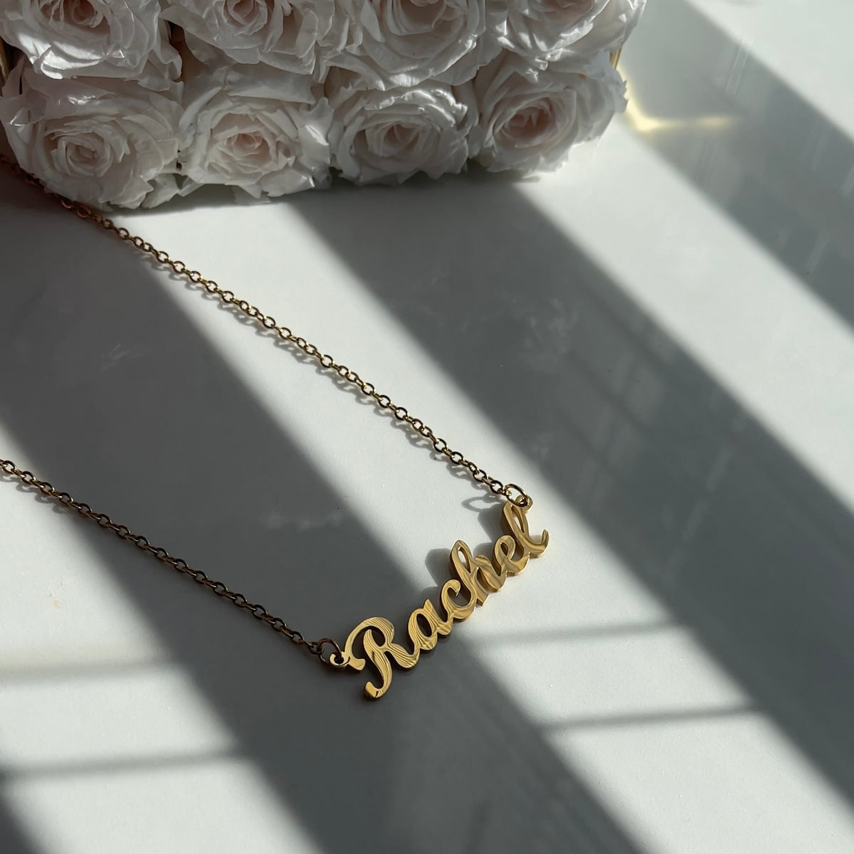 Gold 'Rachel' necklace on a white surface with white roses in the background