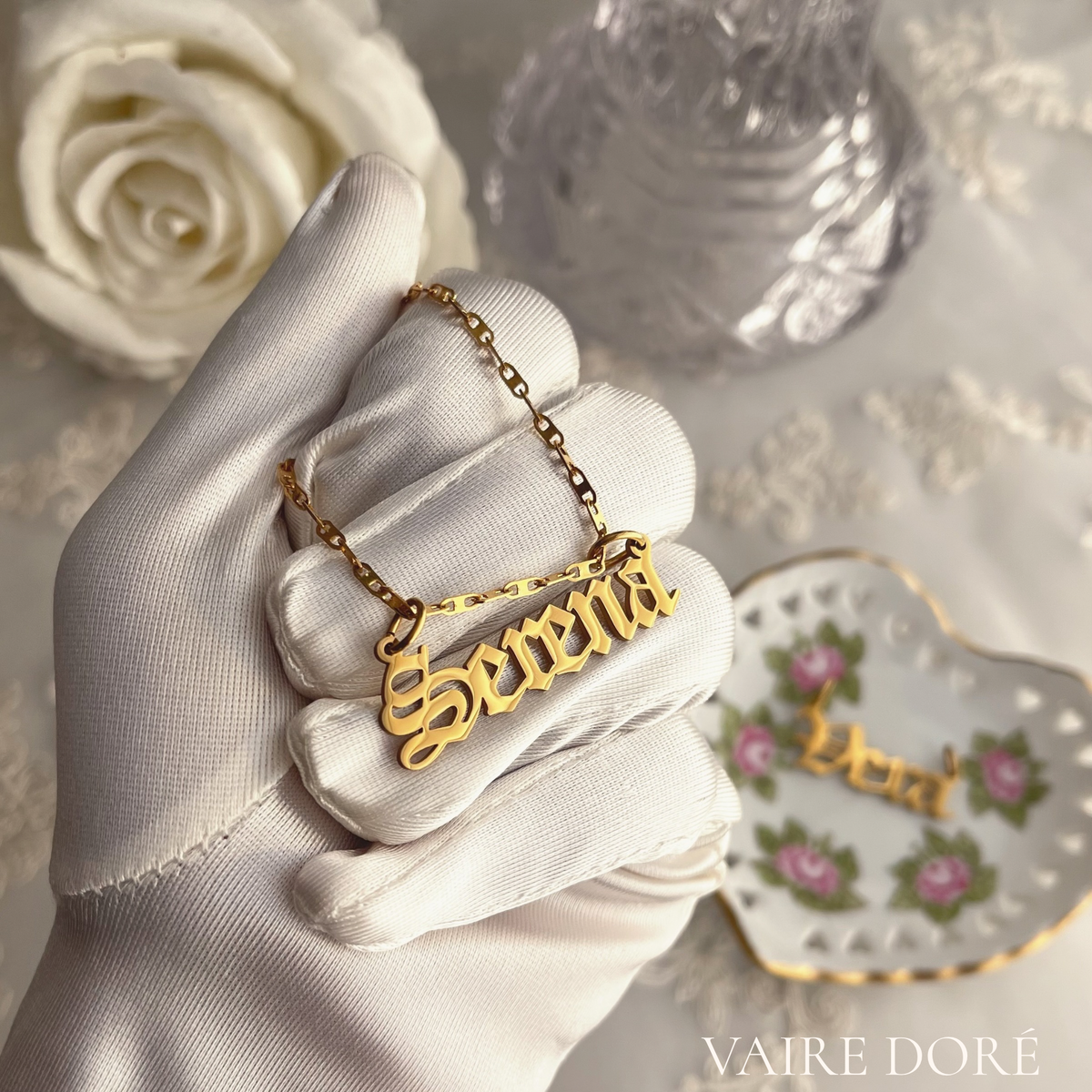 A gold gothic nameplate necklace reading 'Serena' is held on a white satin-gloved hand. The background features a white rose, a textured silver vase, and a heart-shaped floral porcelain dish, with the brand name 'VAIRE DORÉ' in the bottom right corner.