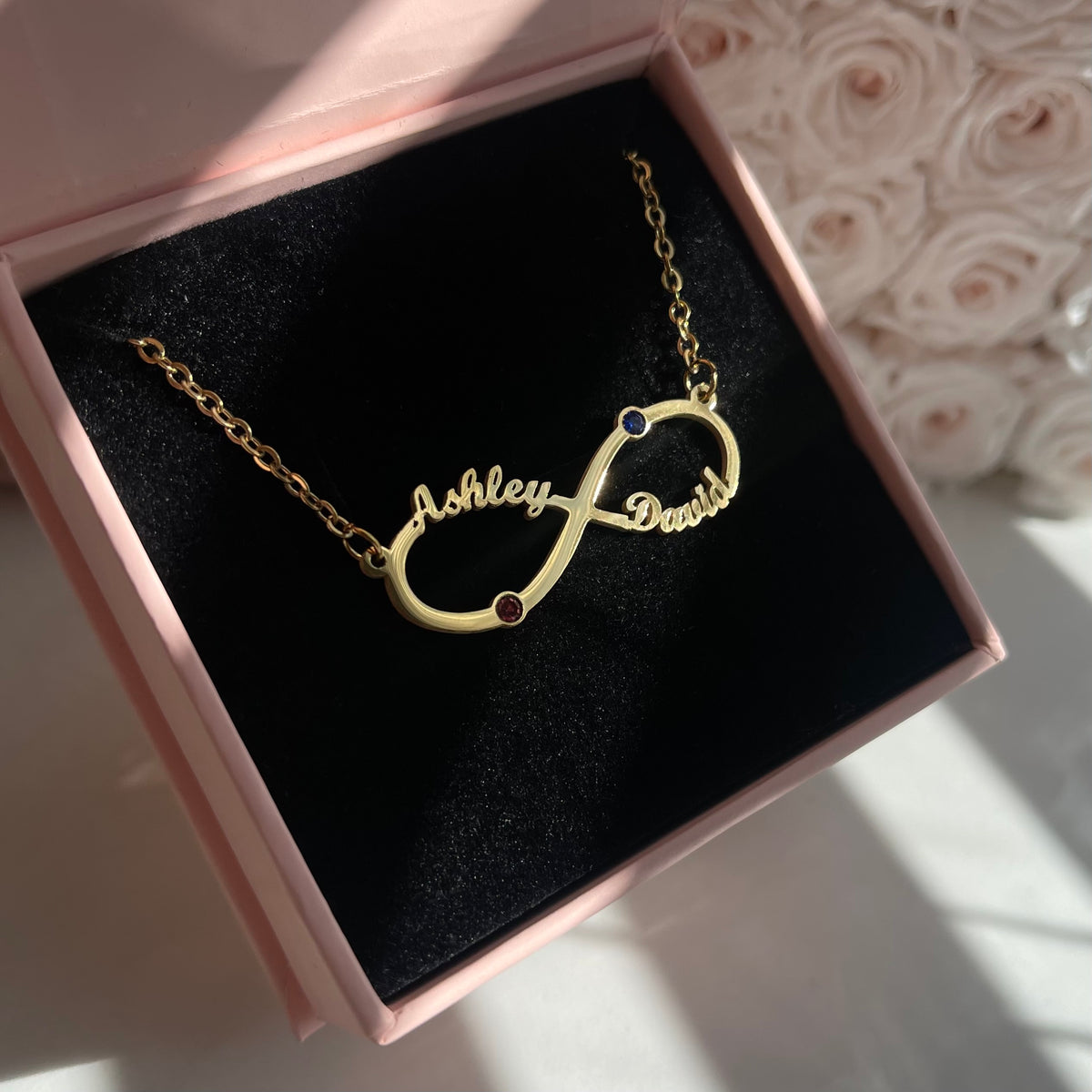 Gold infinity necklace with engraved names in a pink box with floral background