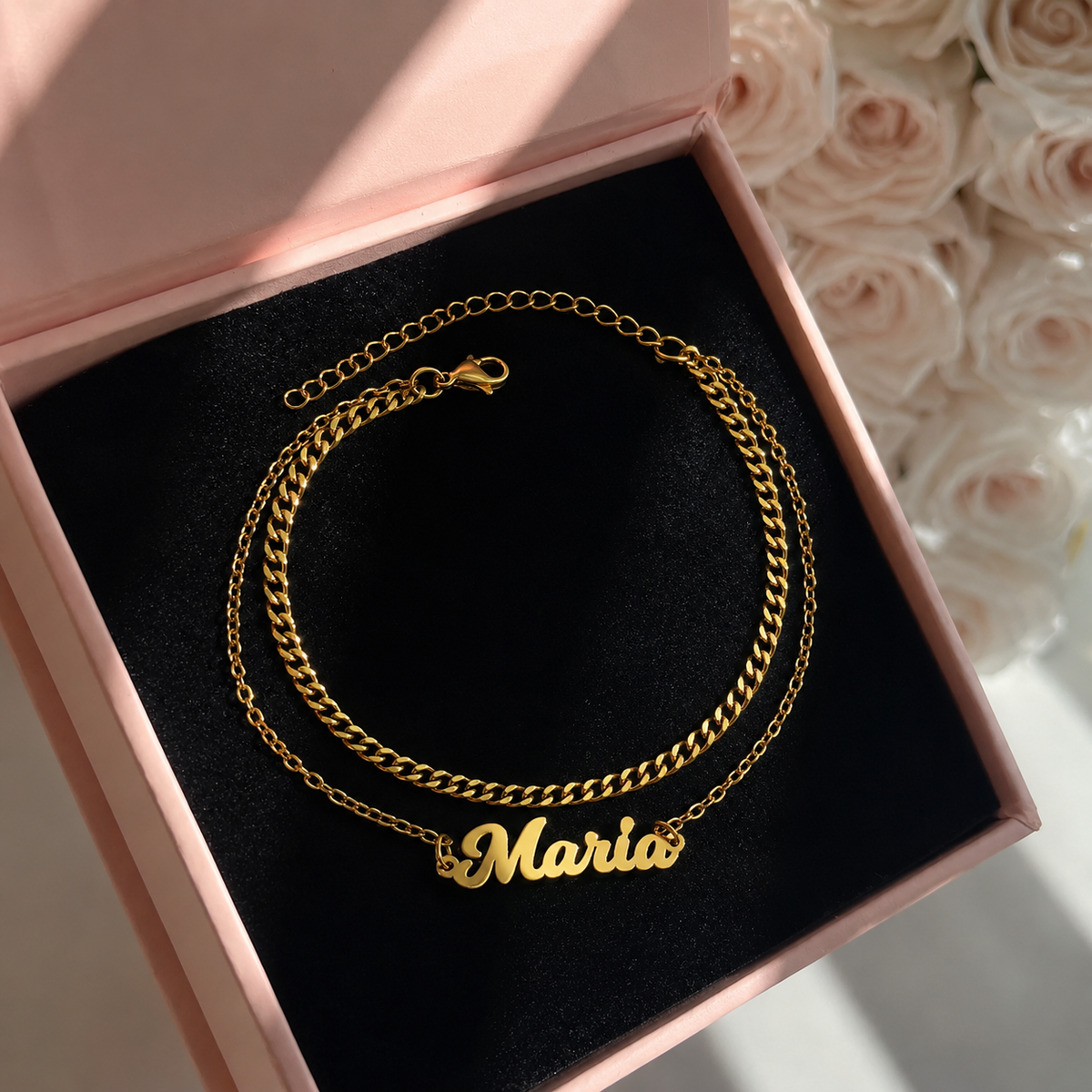 Gold chain necklace with 'Maria' pendant in a pink box with flowers in the background