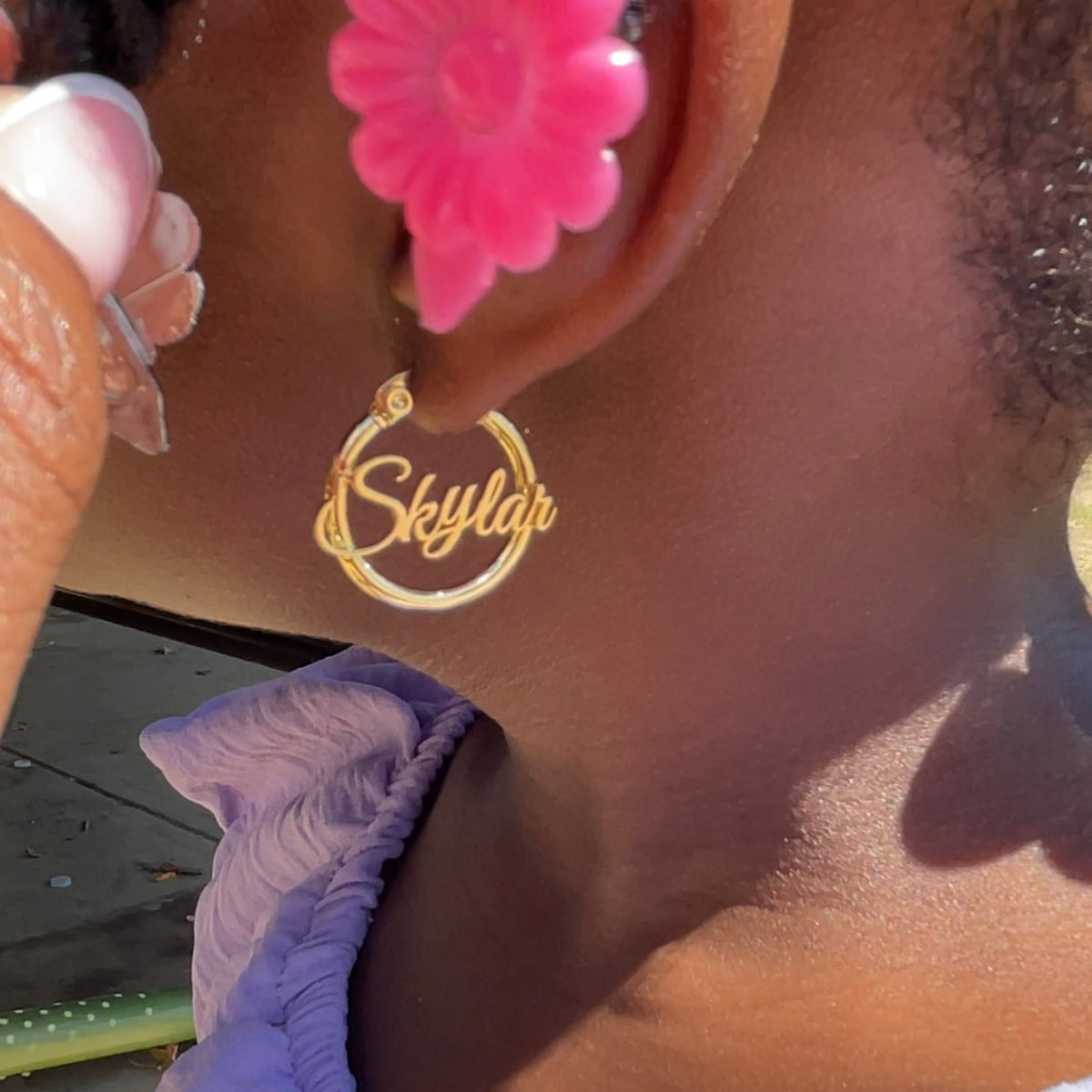 Gold earring with 'Skylar' engraved on it worn by a person with pink flower earrings.