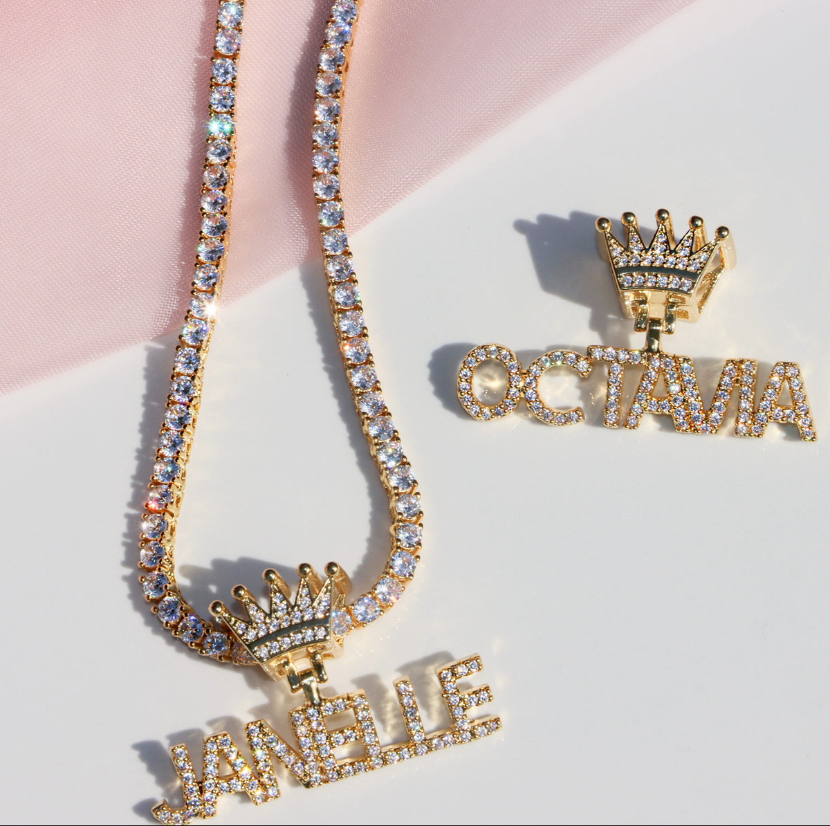 Gold jewelry set with 'JANIE' and 'OCTAVIA' pendants on a pink and white background
