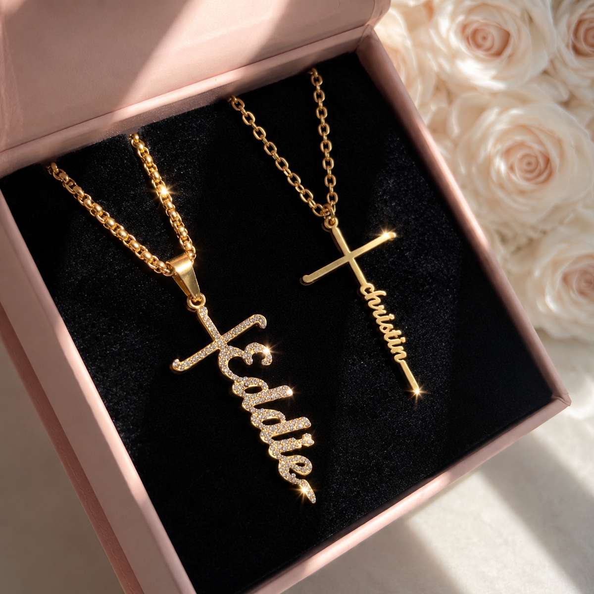 Two gold necklaces with engraved names in a pink box with black interior, surrounded by white roses.