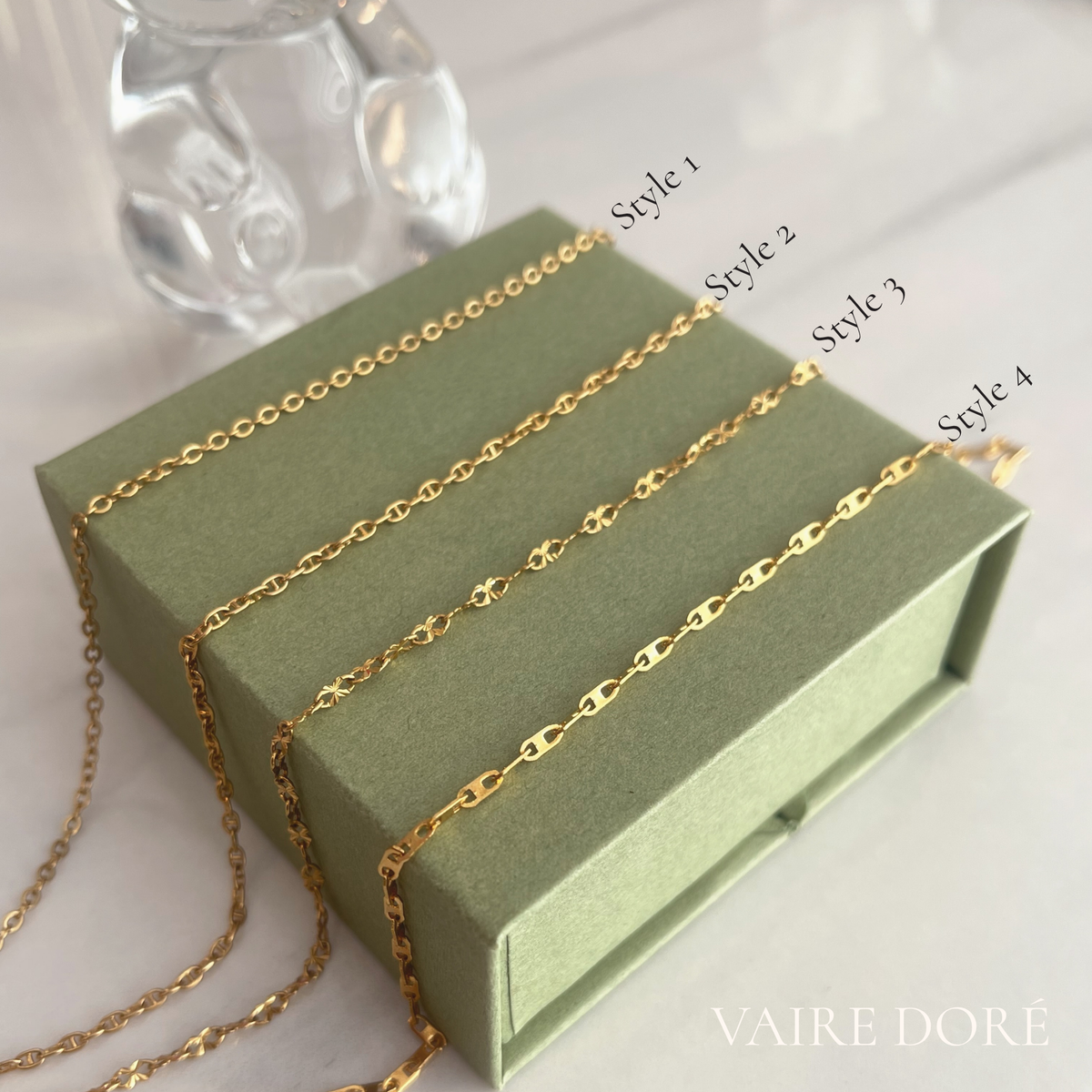 Four gold-toned chain necklaces in different link styles are draped over an olive green jewelry box, with labels reading Style 1 through Style 4. The background is a soft white surface with a blurred glass object behind the box. Four gold-toned chain styles labeled Style 1 through Style 4 draped over an olive green box. Vaire Dore logo on bottom right corner.