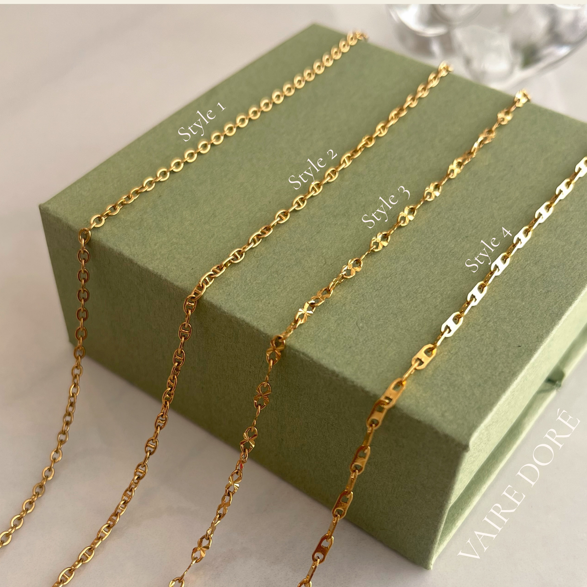 Four gold-toned chain styles labeled Style 1 through Style 4 draped over an olive green box. Vaire Dore logo on bottom right corner.