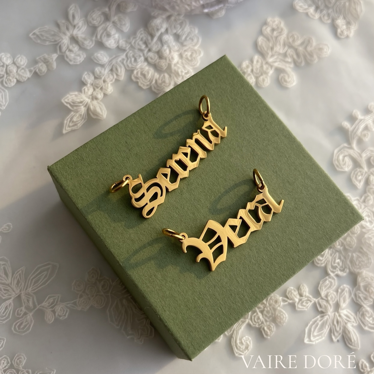 Two gold-toned Gothic pendant necklace spelling 'Serena' and 'Vera' rest on a muted green jewelry box, set against a white lace fabric background.
