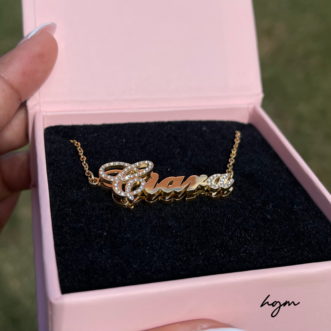 Gold double plated custom name necklace with “Ciara” pendant in script font and crystal accents, displayed in a pink jewelry box outdoors.